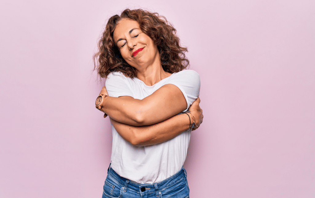 Middle age beautiful woman wearing casual t-shirt standing over isolated pink background hugging herself.